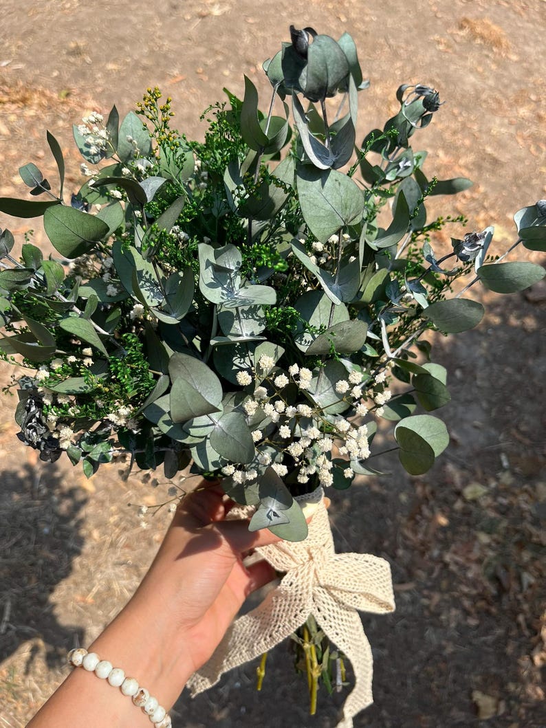 Pode incluir: Um buqu&ecirc; de folhas de eucalipto secas, pequenas flores brancas de gips&oacute;fila e folhagem verde. Os caules est&atilde;o reunidos e amarrados com uma fita de tecido cor de creme de trama aberta, atada num la&ccedil;o decorativo.