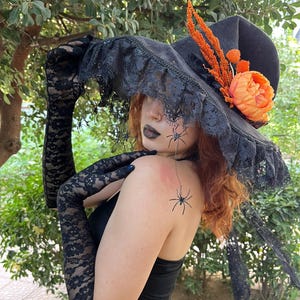 May include: Black witch hat with a wide brim, adorned with black lace trim, orange flowers, and decorative spiders. The hat is paired with long black lace gloves, creating a Halloween or costume theme.