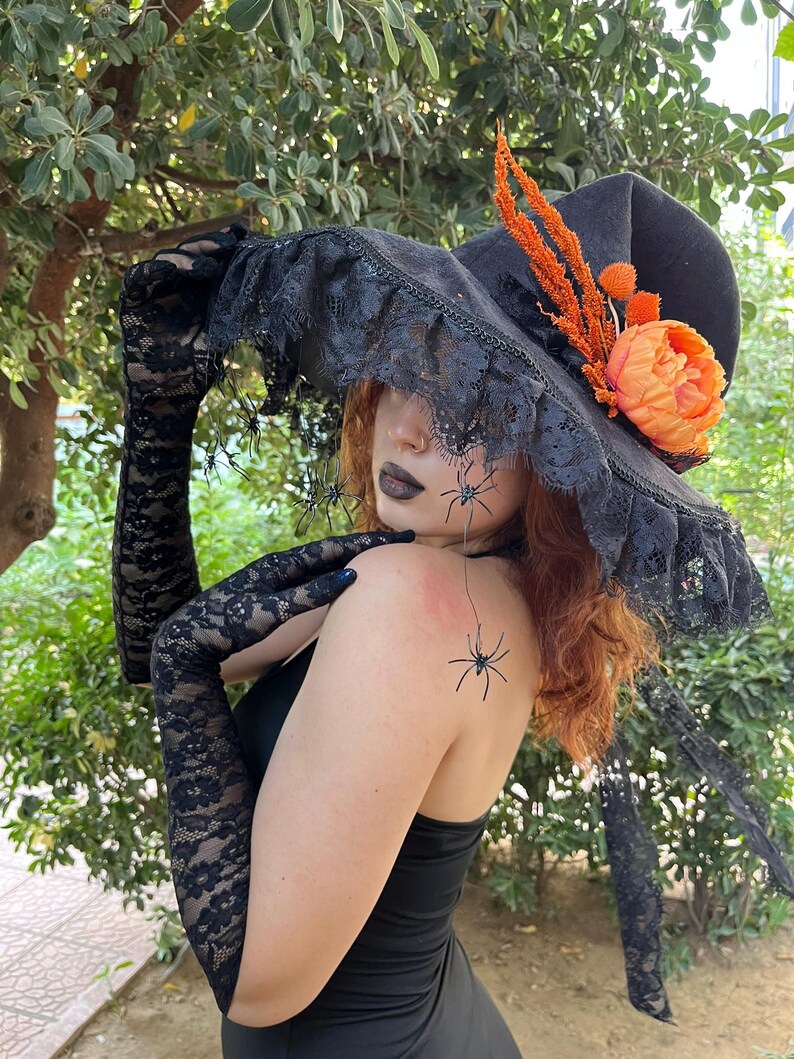 May include: Black witch hat with a wide brim, adorned with black lace trim, orange flowers, and decorative spiders. The hat is paired with long black lace gloves, creating a Halloween or costume theme.