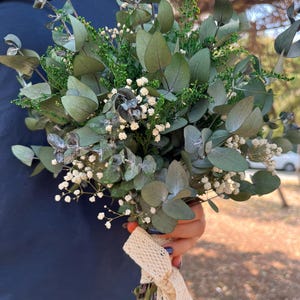 Pode incluir: Um buqu&ecirc; de folhas de eucalipto secas, folhagem verde pequena e min&uacute;sculas flores brancas de gips&oacute;fila. Os caules est&atilde;o reunidos e amarrados com uma fita de renda larga na cor creme, apresentando um delicado padr&atilde;o de losangos.