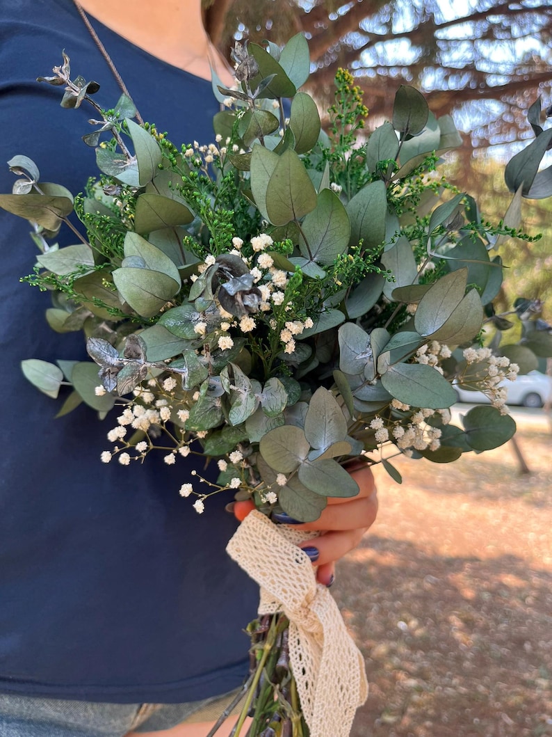 Pode incluir: Um buqu&ecirc; de folhas de eucalipto secas, folhagem verde pequena e min&uacute;sculas flores brancas de gips&oacute;fila. Os caules est&atilde;o reunidos e amarrados com uma fita de renda larga na cor creme, apresentando um delicado padr&atilde;o de losangos.