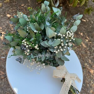 Pode incluir: Um buqu&ecirc; de folhas de eucalipto secas, pequenas flores brancas de gips&oacute;fila e folhagem verde. Os caules est&atilde;o reunidos e presos com uma fita de renda bege de trama aberta, atada num la&ccedil;o na base do arranjo floral.