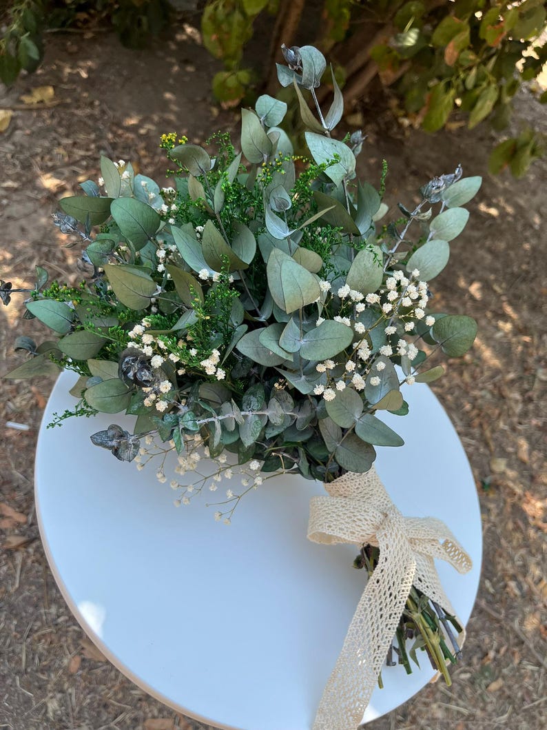 Pode incluir: Um buqu&ecirc; de folhas de eucalipto secas, pequenas flores brancas de gips&oacute;fila e folhagem verde. Os caules est&atilde;o reunidos e presos com uma fita de renda bege de trama aberta, atada num la&ccedil;o na base do arranjo floral.