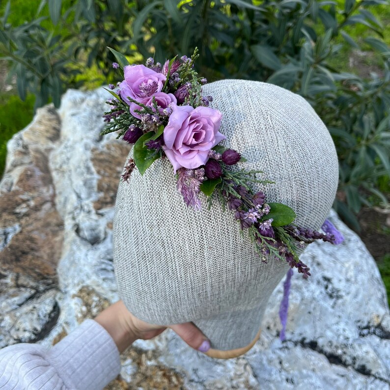 May include: A floral headpiece featuring lavender roses and various purple flowers and greenery. The arrangement is set on a gray head form, with a purple ribbon. The headpiece is designed for special occasions.