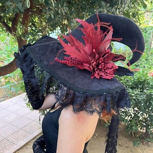 May include: Black witch hat with a curved brim, adorned with a large burgundy flower and feathers. The hat features a lace trim and is part of a costume ensemble. The hat is made of a dark, velvety material.