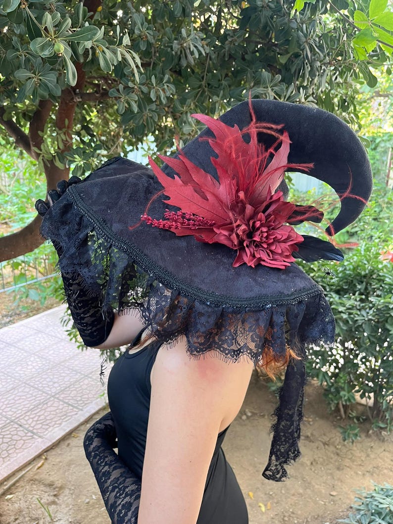May include: Black witch hat with a curved brim, adorned with a large burgundy flower and feathers. The hat features a lace trim and is part of a costume ensemble. The hat is made of a dark, velvety material.