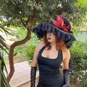 May include: A black halter dress with matching lace gloves. A black hat with a wide brim, adorned with red feathers and black lace trim, completes the ensemble. The outfit is set against a backdrop of green foliage.