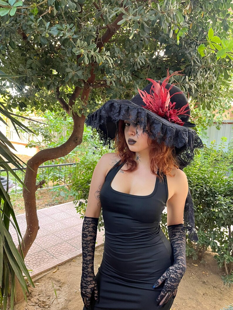 May include: A black halter dress with matching lace gloves. A black hat with a wide brim, adorned with red feathers and black lace trim, completes the ensemble. The outfit is set against a backdrop of green foliage.
