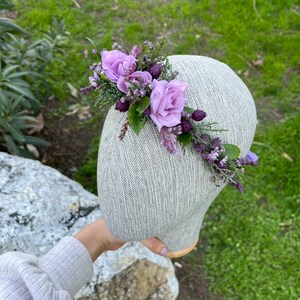 May include: A floral crown featuring lavender roses, sprigs of greenery, and purple accents. The arrangement is set on a neutral-toned head form, showcasing the delicate details of the flowers. Perfect for weddings or special occasions.