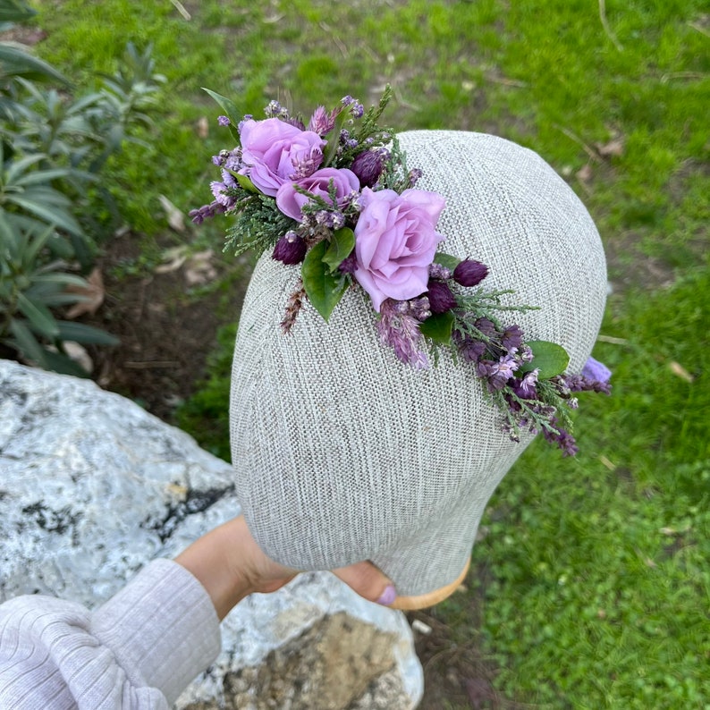 May include: A floral crown featuring lavender roses, sprigs of greenery, and purple accents. The arrangement is set on a neutral-toned head form, showcasing the delicate details of the flowers. Perfect for weddings or special occasions.