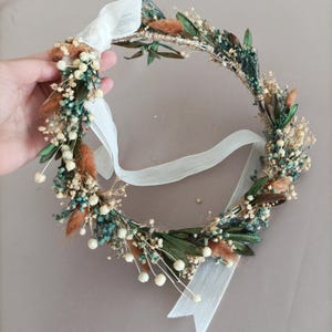 May include: A floral crown featuring a circular design with a variety of dried flowers and greenery. The crown includes small white flowers, teal accents, and brown accents. A white ribbon is attached for tying.