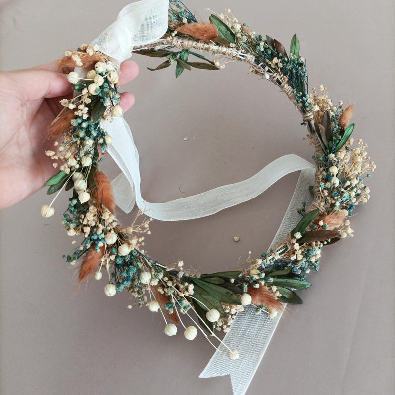 May include: A floral crown featuring a circular design with a variety of dried flowers and greenery. The crown includes small white flowers, teal accents, and brown accents. A white ribbon is attached for tying.