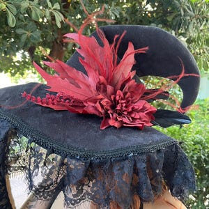 May include: Black witch hat with a wide brim, adorned with a large red flower, red leaves, and black feathers. The brim is trimmed with black lace. The hat is set against a backdrop of green foliage.