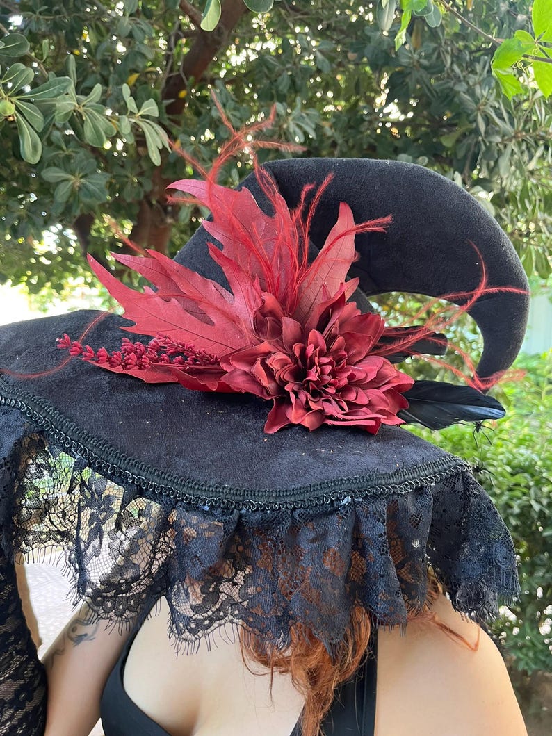 May include: Black witch hat with a wide brim, adorned with a large red flower, red leaves, and black feathers. The brim is trimmed with black lace. The hat is set against a backdrop of green foliage.