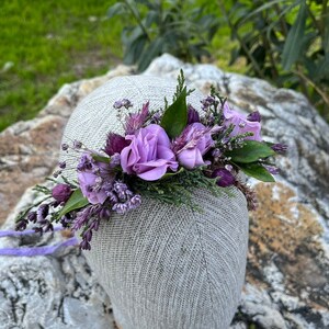 May include: A floral crown featuring lavender and purple flowers, including roses, set against a backdrop of green foliage. The crown is attached to a gray head form, with a lavender ribbon extending from the base. Perfect for weddings or special events.
