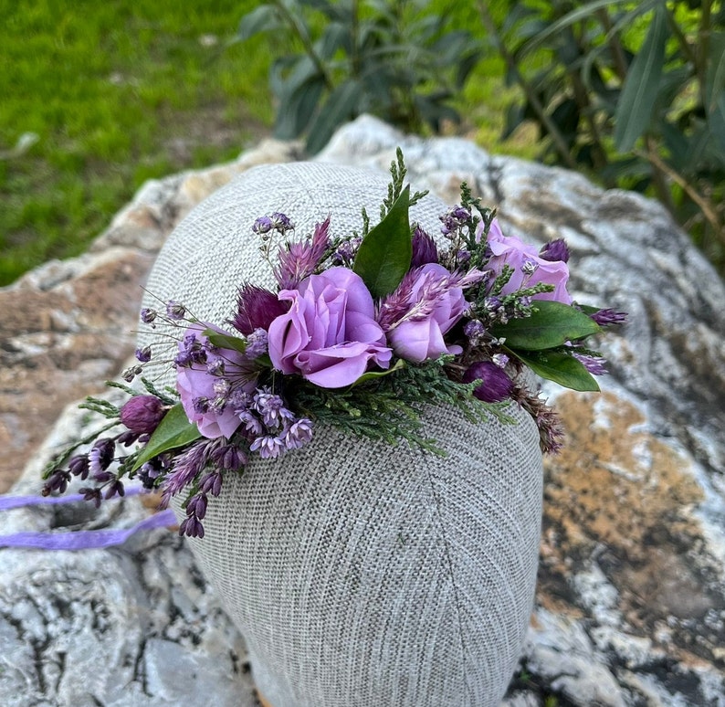 May include: A floral crown featuring lavender and purple flowers, including roses, set against a backdrop of green foliage. The crown is attached to a gray head form, with a lavender ribbon extending from the base. Perfect for weddings or special events.