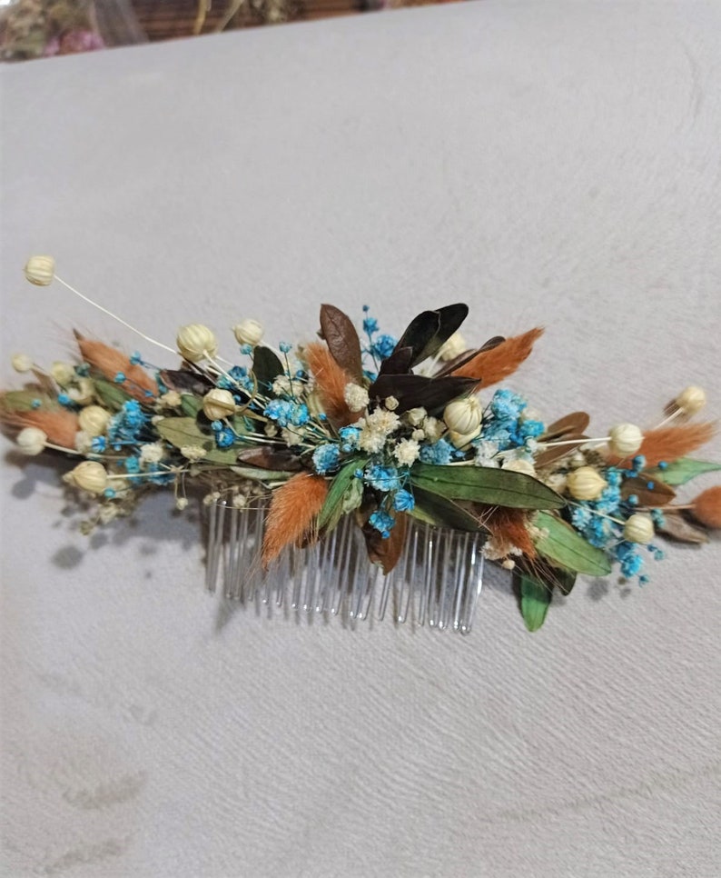 May include: A floral hair comb featuring a variety of dried flowers in shades of brown, orange, blue, and green. The comb has a clear plastic base with multiple teeth, and the flowers are arranged in a decorative pattern.
