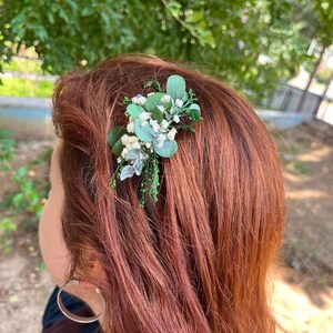 Pode incluir: Um acess&oacute;rio de cabelo com um grupo de pequenas flores brancas de gips&oacute;fila e folhas verdes de eucalipto. O arranjo floral est&aacute; preso a uma presilha de cabelo, exibindo elementos bot&acirc;nicos naturais com flores brancas.