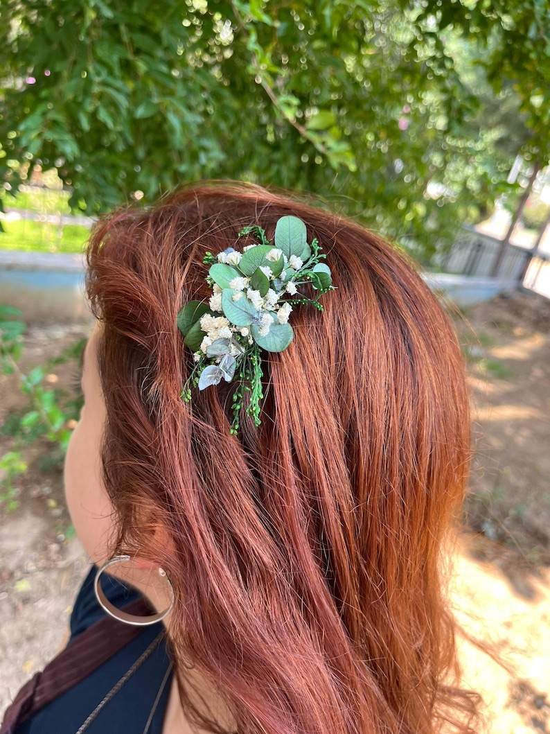 Pode incluir: Um acess&oacute;rio de cabelo com um grupo de pequenas flores brancas de gips&oacute;fila e folhas verdes de eucalipto. O arranjo floral est&aacute; preso a uma presilha de cabelo, exibindo elementos bot&acirc;nicos naturais com flores brancas.