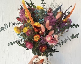 Pode incluir: Um vibrante buquê de flores secas com flores amarelas, laranjas, rosas, roxas e azuis misturadas com caules de eucalipto verdes. Os caules são reunidos e amarrados com uma fita de renda branca, criando um arranjo colorido.