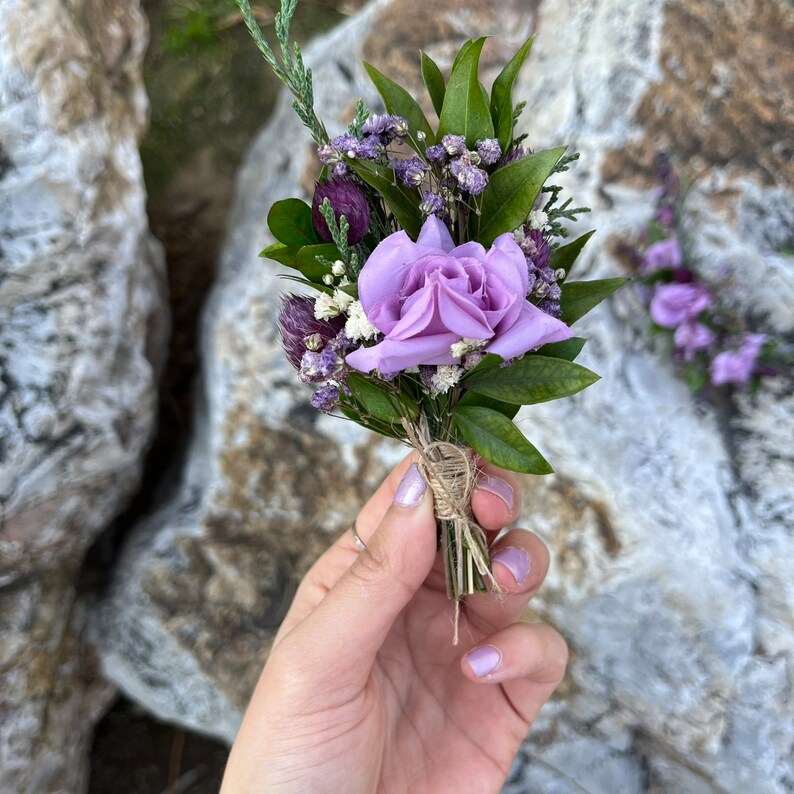May include: A small, handcrafted floral arrangement featuring a light purple rose as the focal point, surrounded by various purple and white accent flowers and green leaves. The stems are bound with twine, creating a rustic and natural aesthetic.
