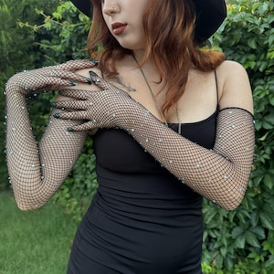 May include: A person wearing a black sleeveless dress and black fishnet gloves with rhinestones. The gloves are long and reach to the elbows.