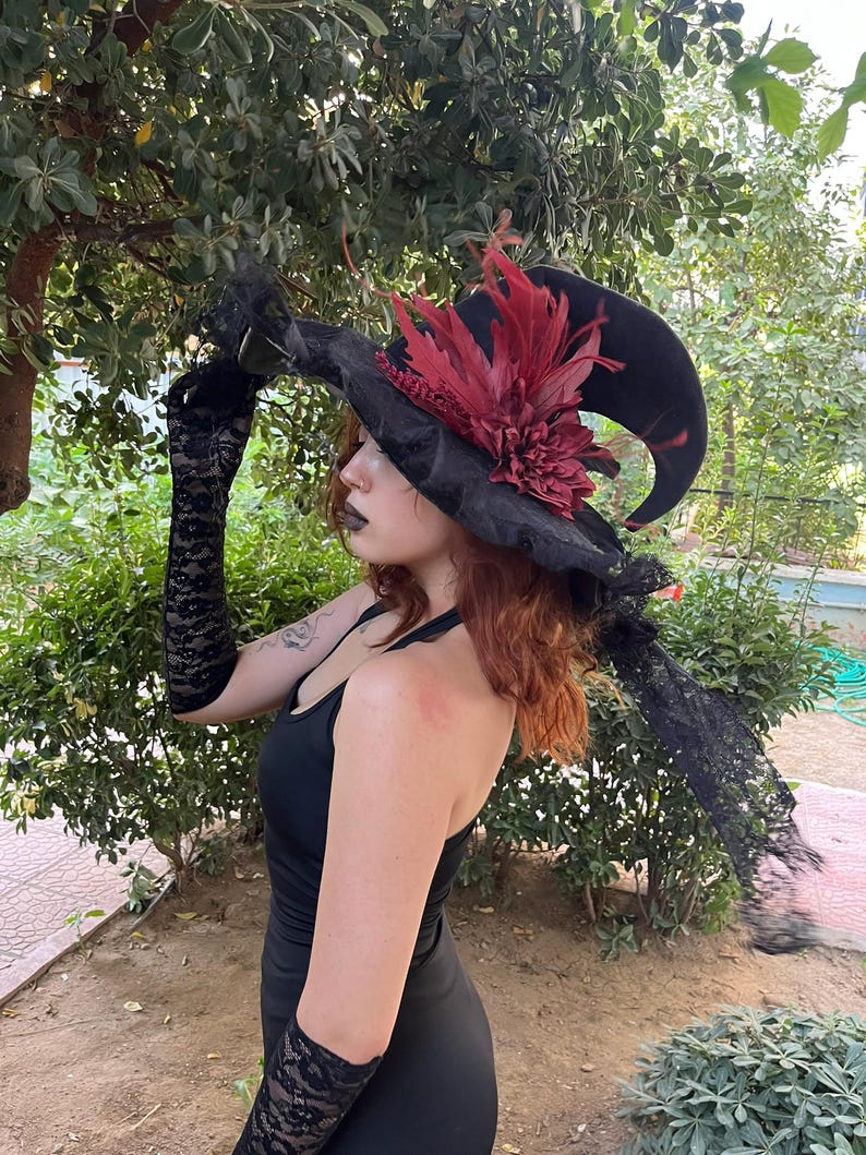 May include: A black witch hat with a wide brim, adorned with a large burgundy flower and feathers. The hat has a curved, pointed tip. Black lace gloves and a black dress complete the gothic ensemble, set against a backdrop of green foliage.
