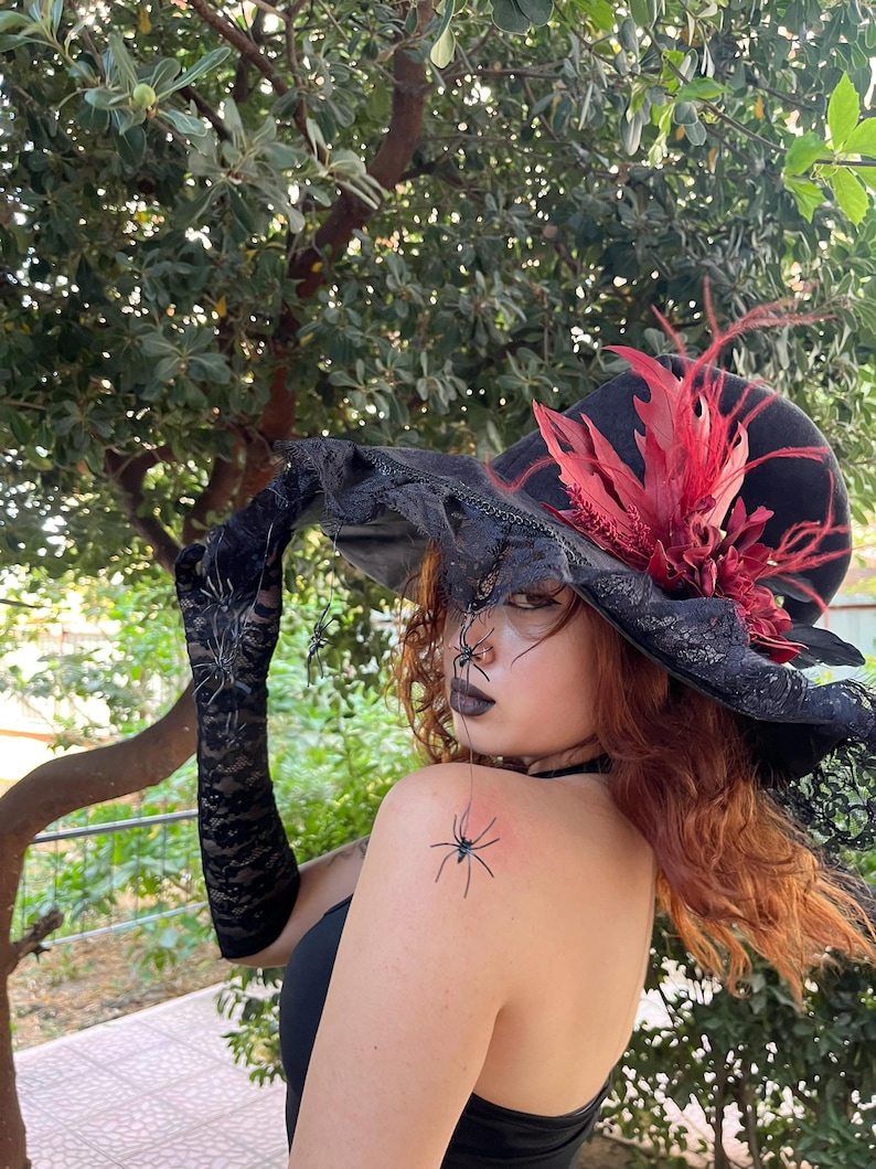 May include: A black witch hat with a wide brim, adorned with red feathers and lace trim. The hat is part of a Halloween costume, with black lace gloves and spider decorations.