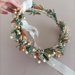 A delicate floral crown made with dried flowers in shades of green, white, and brown. The crown is adorned with small white flowers, dried leaves, and sprigs of dried grasses. A white ribbon is attached to the crown.