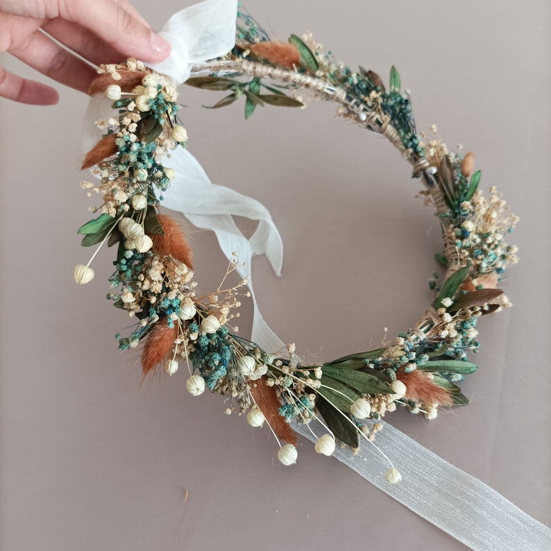 A delicate floral crown made with dried flowers in shades of green, white, and brown. The crown is adorned with small white flowers, dried leaves, and sprigs of dried grasses. A white ribbon is attached to the crown.