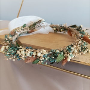 May include: A floral crown featuring a variety of dried flowers in shades of teal, cream, and brown. The crown is constructed on a natural twine base and includes accents of green leaves and fluffy brown accents. White ribbon ties the crown together.