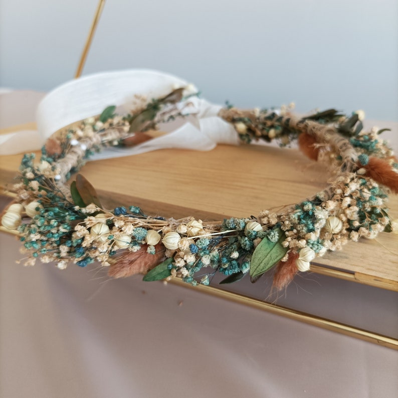 May include: A floral crown featuring a variety of dried flowers in shades of teal, cream, and brown. The crown is constructed on a natural twine base and includes accents of green leaves and fluffy brown accents. White ribbon ties the crown together.