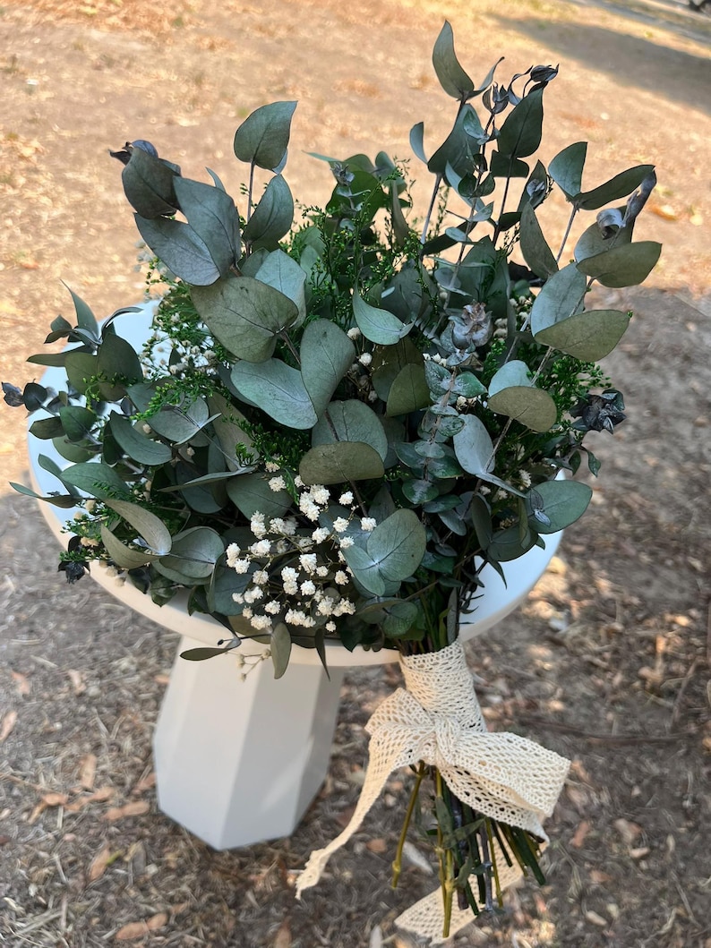 Pode incluir: Um buqu&ecirc; de folhas de eucalipto secas e pequenas flores brancas de gips&oacute;fila, amarrado com uma fita de renda na cor creme. Os caules est&atilde;o reunidos na base e a folhagem apresenta tons verdes e cinzentos suaves em todo o arranjo.