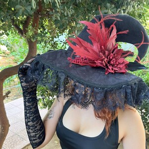 May include: A black witch hat with a wide brim, adorned with a red floral arrangement and feathers. The hat features a lace trim and is part of a costume ensemble. The image is set against a backdrop of green foliage.