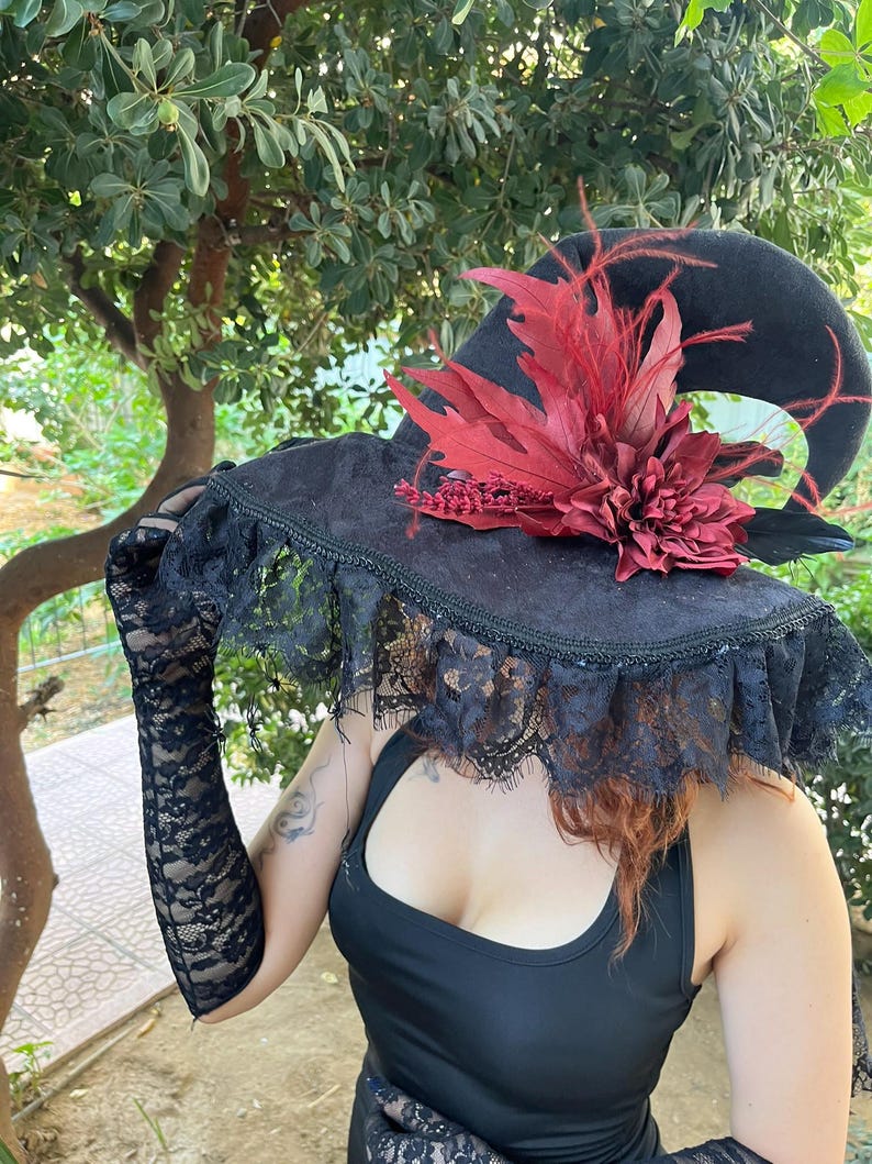 May include: A black witch hat with a wide brim, adorned with a red floral arrangement and feathers. The hat features a lace trim and is part of a costume ensemble. The image is set against a backdrop of green foliage.