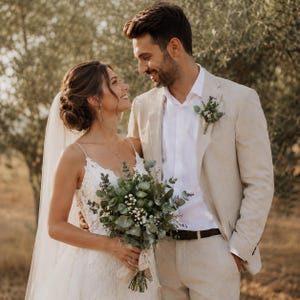 Pode incluir: Um vestido de noiva de renda branca com al&ccedil;as finas e um v&eacute;u longo, combinado com um palet&oacute; bege, uma camisa branca e um cinto escuro. O conjunto inclui um buqu&ecirc; de eucalipto e gips&oacute;fila e uma flor de lapela floral correspondente.