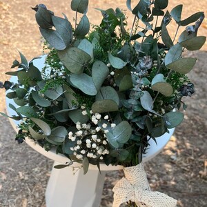 Pode incluir: Um buqu&ecirc; de ramos de eucalipto secos com folhas arredondadas de tom verde suave, intercaladas com pequenos cachos de delicadas flores brancas de gips&oacute;fila e folhagem verde fina. Os caules est&atilde;o reunidos e atados com uma fita de renda cor de creme.