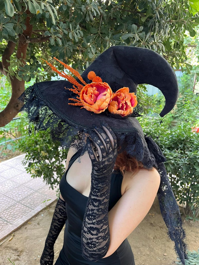 May include: A black witch hat with a wide brim, adorned with orange and pink flowers and lace trim. The hat has a pointed crown and is decorated with orange sprigs. The hat is a costume accessory.