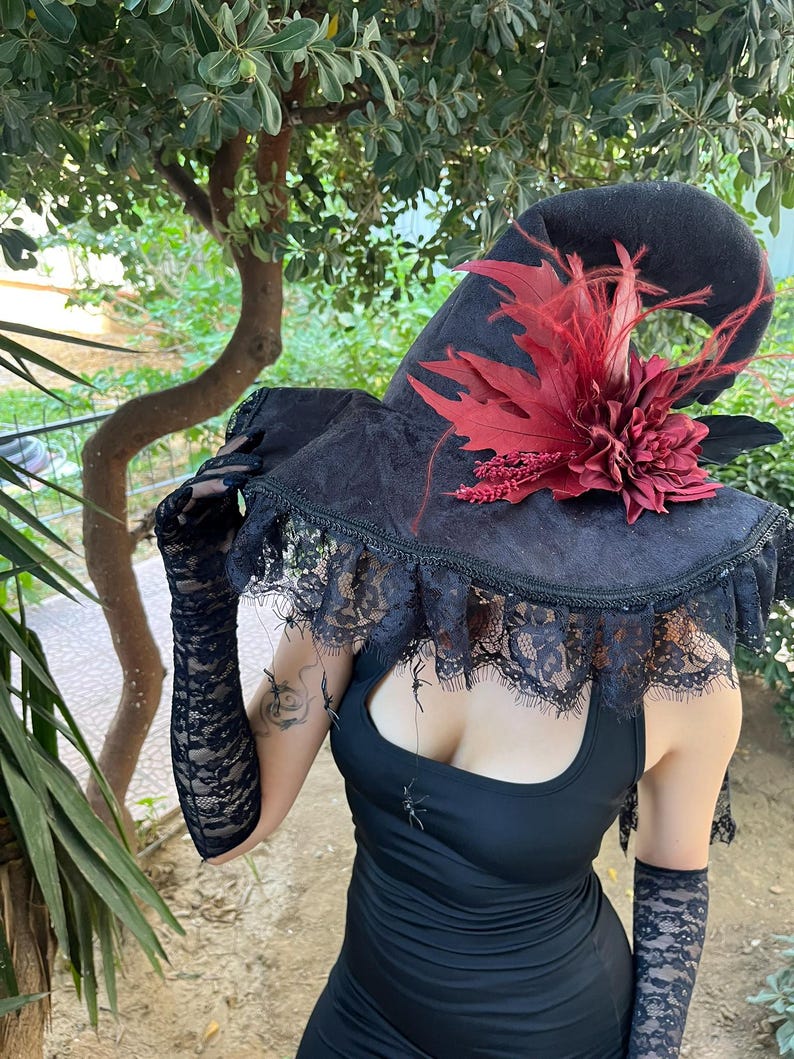May include: A black witch hat with a wide brim, adorned with red leaves and a flower. The hat has a lace trim and is paired with black lace gloves. The overall theme is gothic and Halloween.