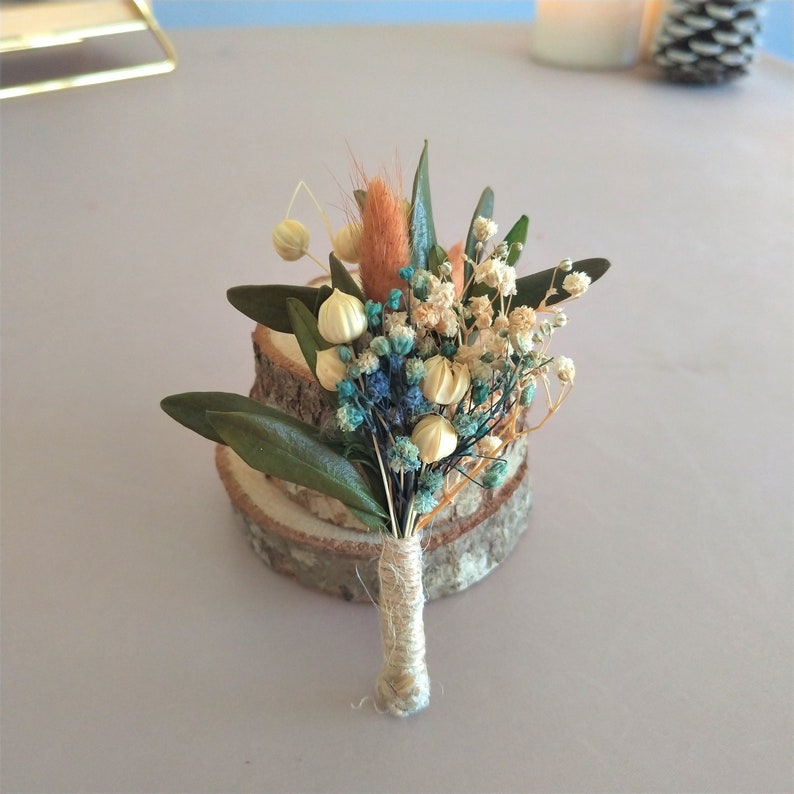 May include: A small, dried flower boutonniere with a variety of textures and colors. The arrangement includes blue, white, and orange flowers, with green leaves. The stems are wrapped in twine, and the boutonniere rests on a wooden slice.