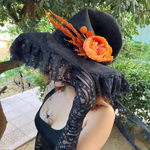 May include: Black witch hat with a wide brim, adorned with orange flowers and accents. The hat features a lace trim and is decorated with small plastic spiders, perfect for Halloween or costume parties.