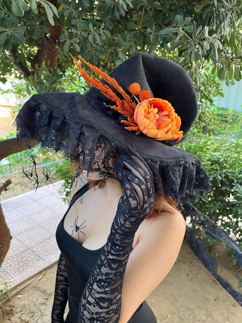 May include: Black witch hat with a wide brim, adorned with orange flowers and accents. The hat features a lace trim and is decorated with small plastic spiders, perfect for Halloween or costume parties.