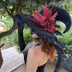 May include: A black witch hat with a curved brim, adorned with a large burgundy flower and feathers. The hat features a lace trim and is accessorized with black lace gloves and decorative spiders, perfect for Halloween or costume events.