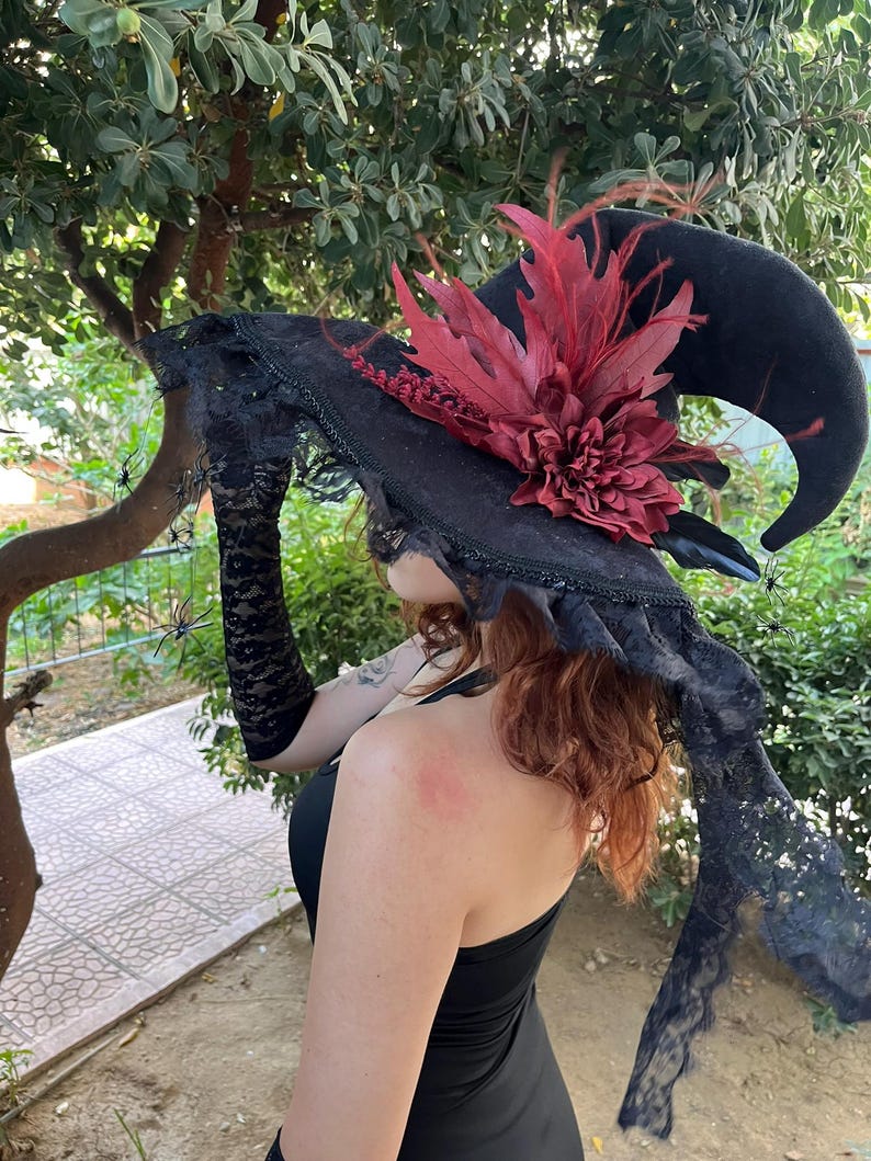May include: A black witch hat with a curved brim, adorned with a large burgundy flower and feathers. The hat features a lace trim and is accessorized with black lace gloves and decorative spiders, perfect for Halloween or costume events.
