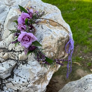 May include: A delicate floral crown featuring lavender roses, sprigs of purple flowers, and green leaves. The crown is set on a thin, brown wire base and has a purple ribbon tie. The arrangement is placed on a large, textured rock.