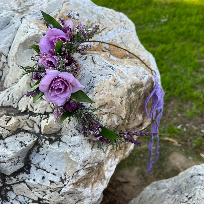 May include: A delicate floral crown featuring lavender roses, sprigs of purple flowers, and green leaves. The crown is set on a thin, brown wire base and has a purple ribbon tie. The arrangement is placed on a large, textured rock.
