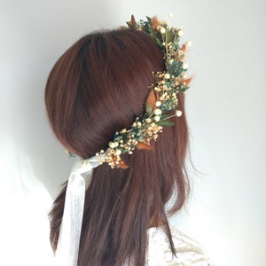 May include: A floral crown featuring a variety of dried flowers and greenery. The crown includes small white flowers, teal accents, and brown leaves. White ribbons are tied at the back. The crown is worn on brown hair.