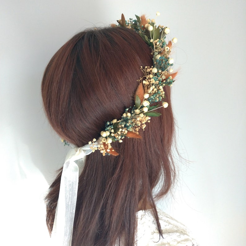 May include: A floral crown featuring a variety of dried flowers and greenery. The crown includes small white flowers, teal accents, and brown leaves. White ribbons are tied at the back. The crown is worn on brown hair.