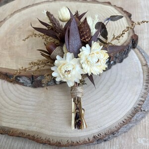 May include: A boutonniere featuring dried cream-colored strawflowers, dark brown eucalyptus leaves, a small acorn, and textured grasses. The stems are bundled together and wrapped with a thin, natural-colored twine to secure the arrangement.