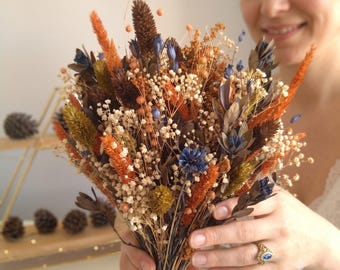 Pode incluir: Um buquê de flores secas com caules laranja, castanhos e azuis misturados com gipsófila branca. O arranjo inclui várias texturas como ervas altas, pequenas flores azuis e vagens castanhas escuras, todas atadas na base.